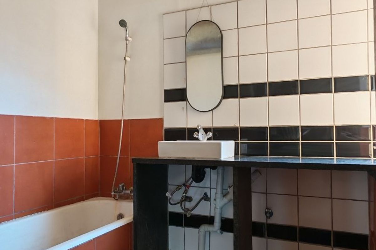 A dated bathroom featuring a bathtub, handheld shower, and a wall-mounted basin with exposed plumbing and decorative tiled walls.