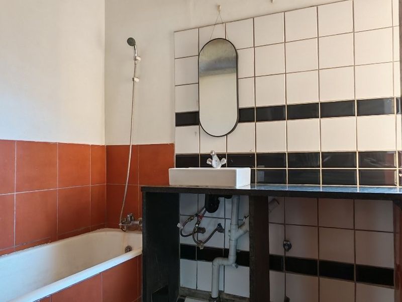 A dated bathroom featuring a bathtub, handheld shower, and a wall-mounted basin with exposed plumbing and decorative tiled walls.