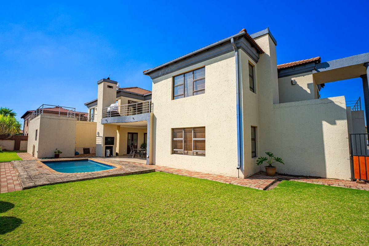 Rear view of the property featuring a swimming pool, landscaped lawn, covered patio with built-in braai, and an upper-level balcony.