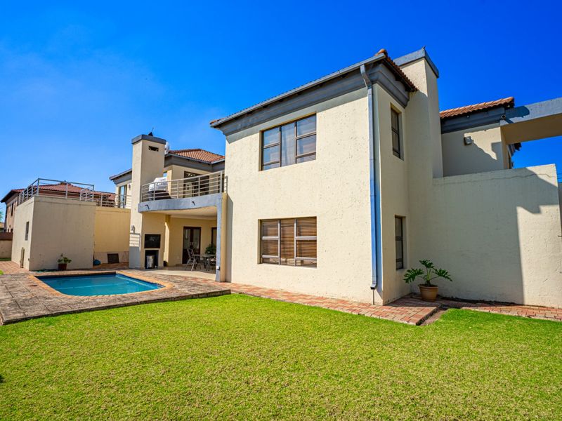 Rear view of the property featuring a swimming pool, landscaped lawn, covered patio with built-in braai, and an upper-level balcony.