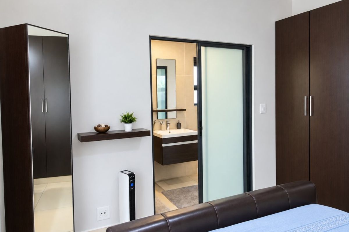 A modern bedroom featuring dark wood built-in cupboards, a sliding door leading to an en-suite bathroom, and neutral tiled flooring.