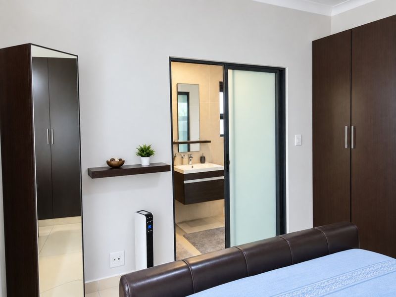 A modern bedroom featuring dark wood built-in cupboards, a sliding door leading to an en-suite bathroom, and neutral tiled flooring.