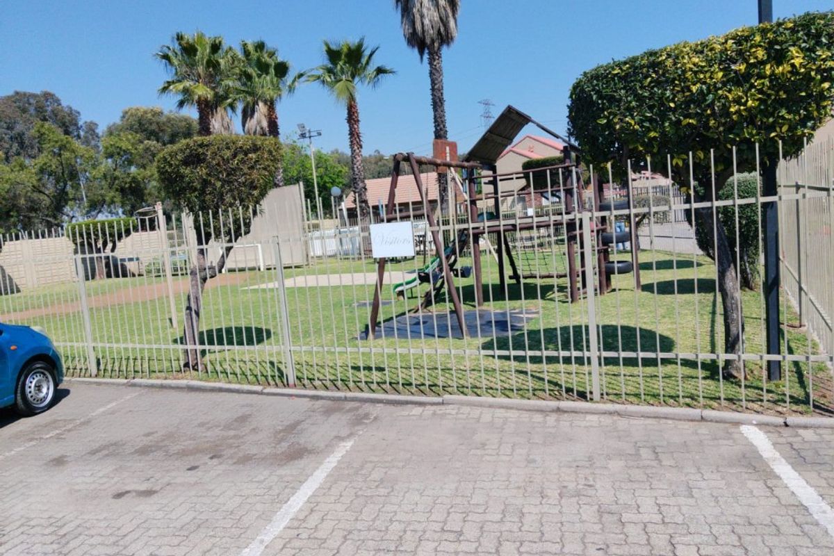 A communal outdoor area featuring a children's playground, manicured lawn, and secure metal perimeter fencing adjacent to a paved parking lot.