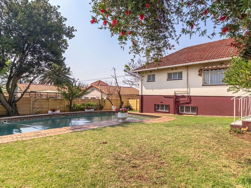 A spacious backyard featuring a swimming pool, brick-paved surround, and established garden area adjacent to the rear elevation of the house.