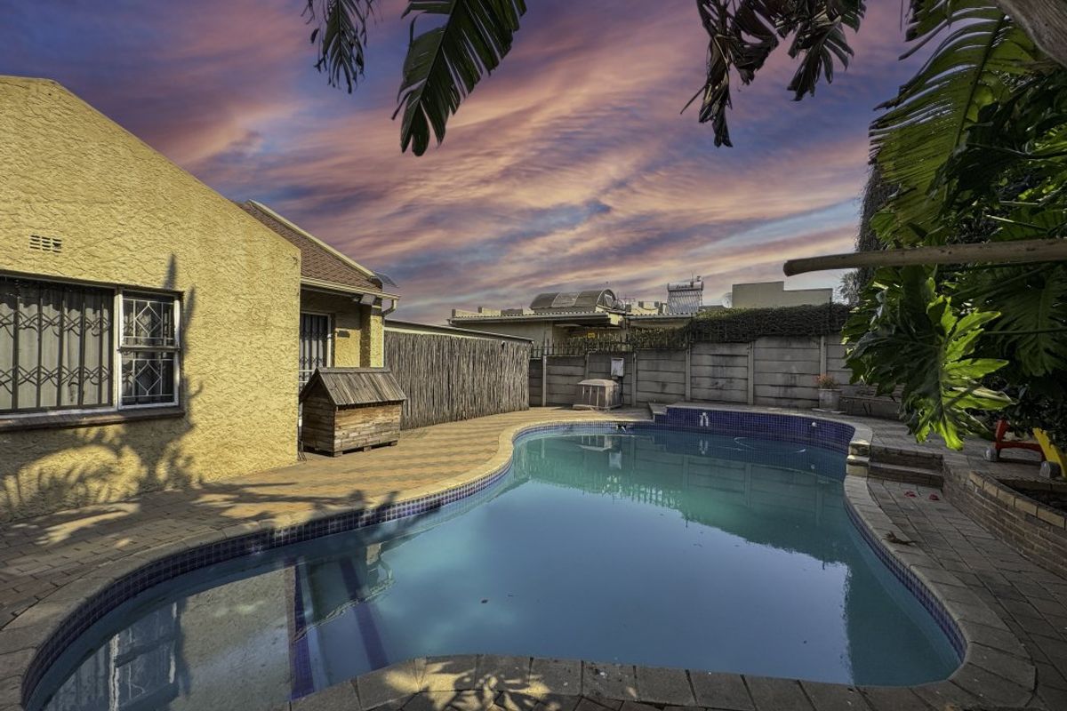 Outdoor pool area featuring a paved patio, swimming pool, and perimeter walling, set against a sunset sky.