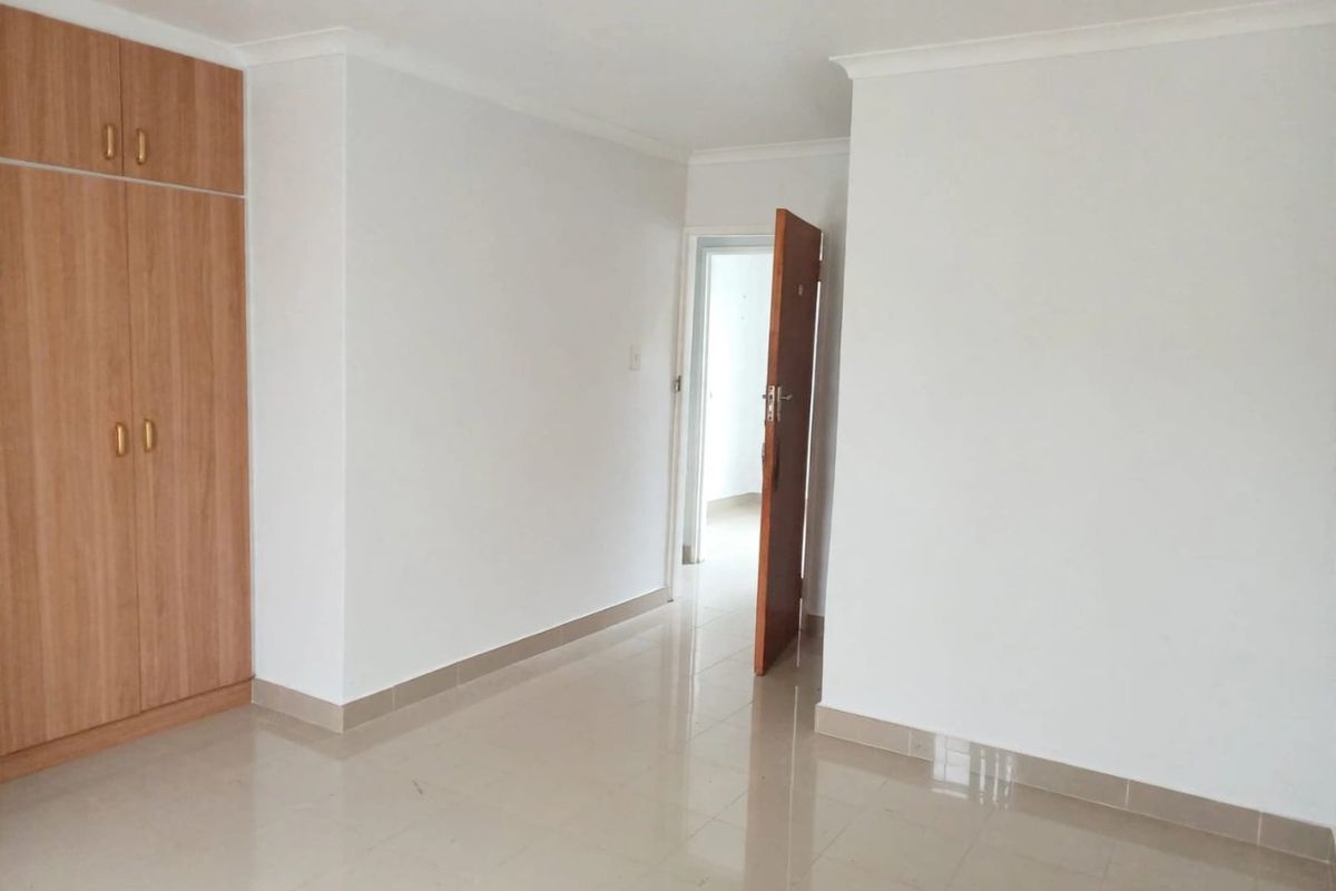 A clean, empty bedroom featuring neutral tiled flooring, white walls, and a built-in wooden wardrobe with overhead storage.