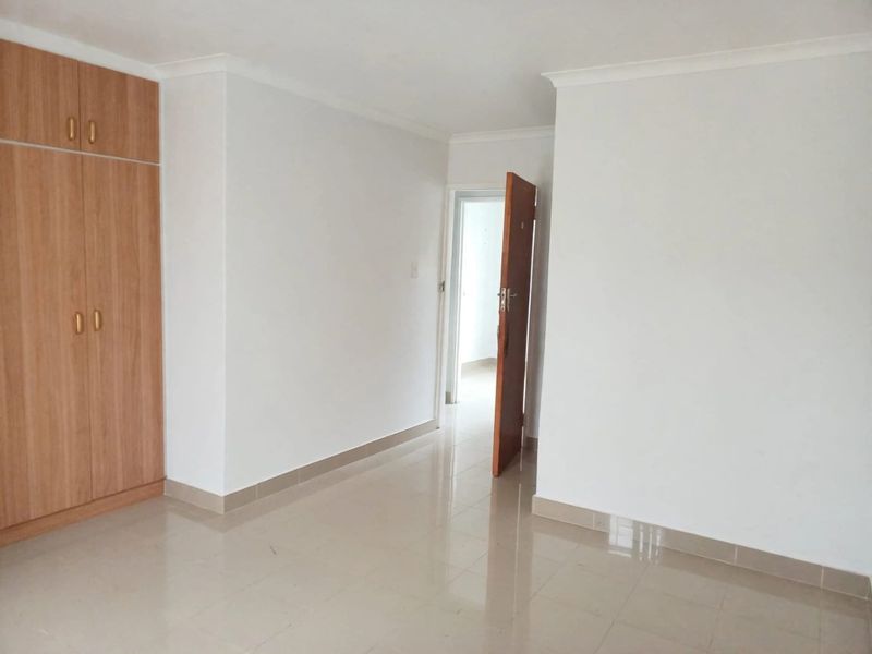 A clean, empty bedroom featuring neutral tiled flooring, white walls, and a built-in wooden wardrobe with overhead storage.