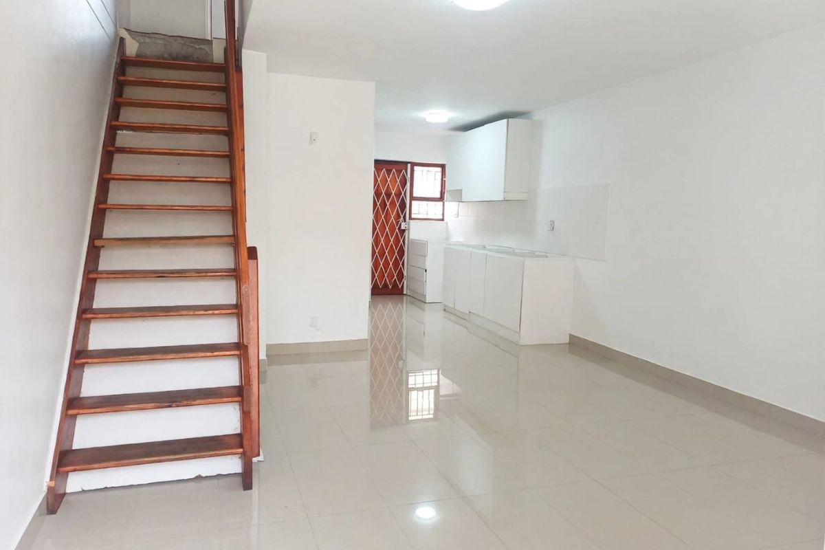 An open-plan living and kitchen area featuring tiled floors, a wooden staircase, and white built-in kitchen cabinetry.