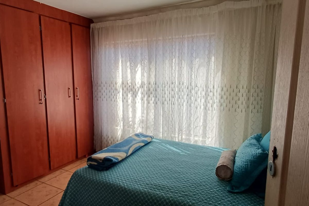 A bedroom featuring built-in wooden cupboards and tiled flooring, with natural light entering through a window covered by sheer curtains.