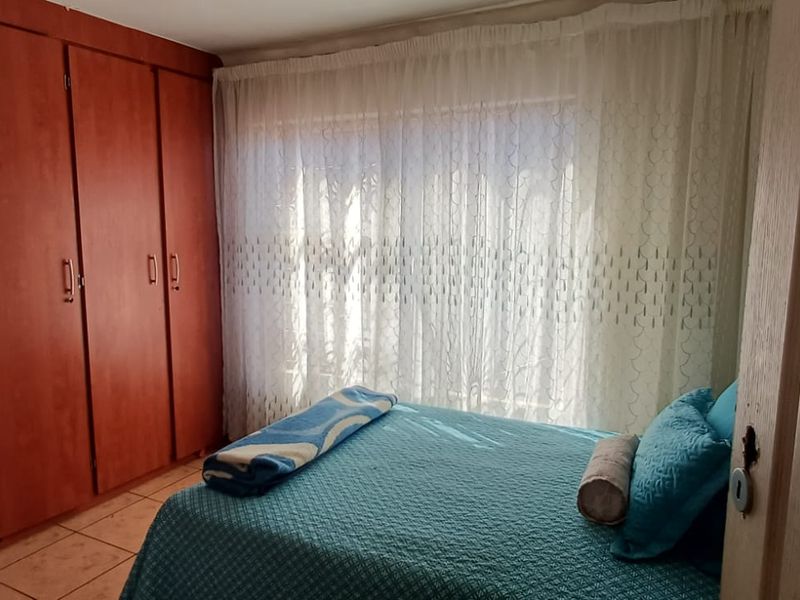 A bedroom featuring built-in wooden cupboards and tiled flooring, with natural light entering through a window covered by sheer curtains.