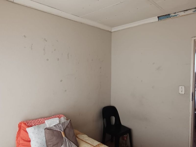 A bedroom interior featuring plain walls with visible surface wear and damaged ceiling trim, with an adjacent view into a bathroom.
