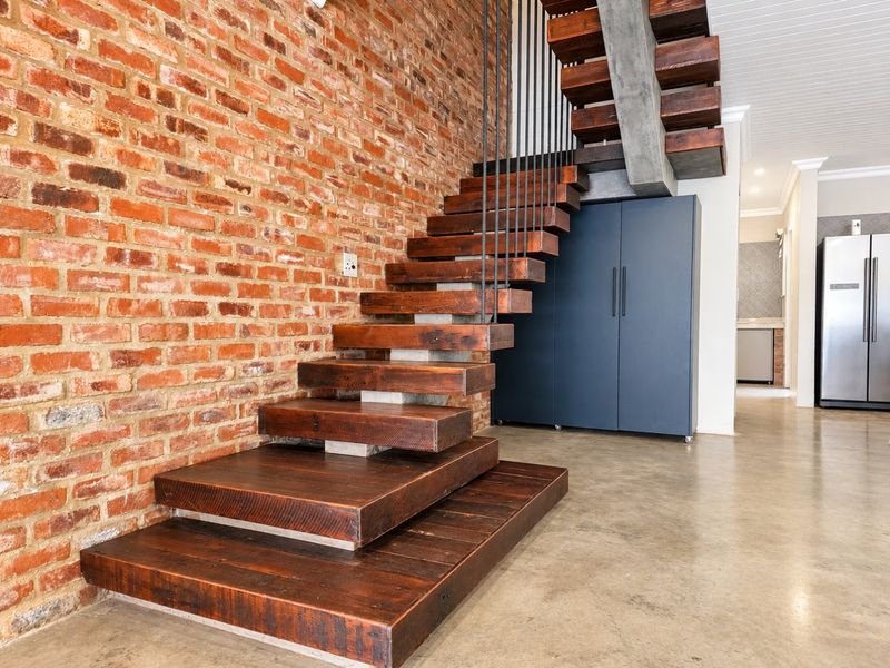 Modern hallway featuring a striking timber staircase with metal balustrade against an exposed brick wall and polished concrete flooring.