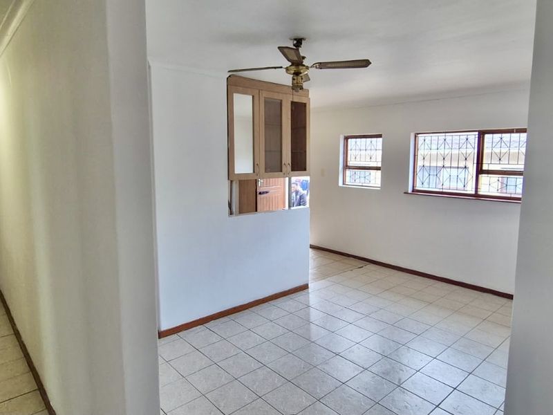 An empty living area featuring tiled flooring, a ceiling fan, and security bars on the windows, with a view into an adjacent hallway.