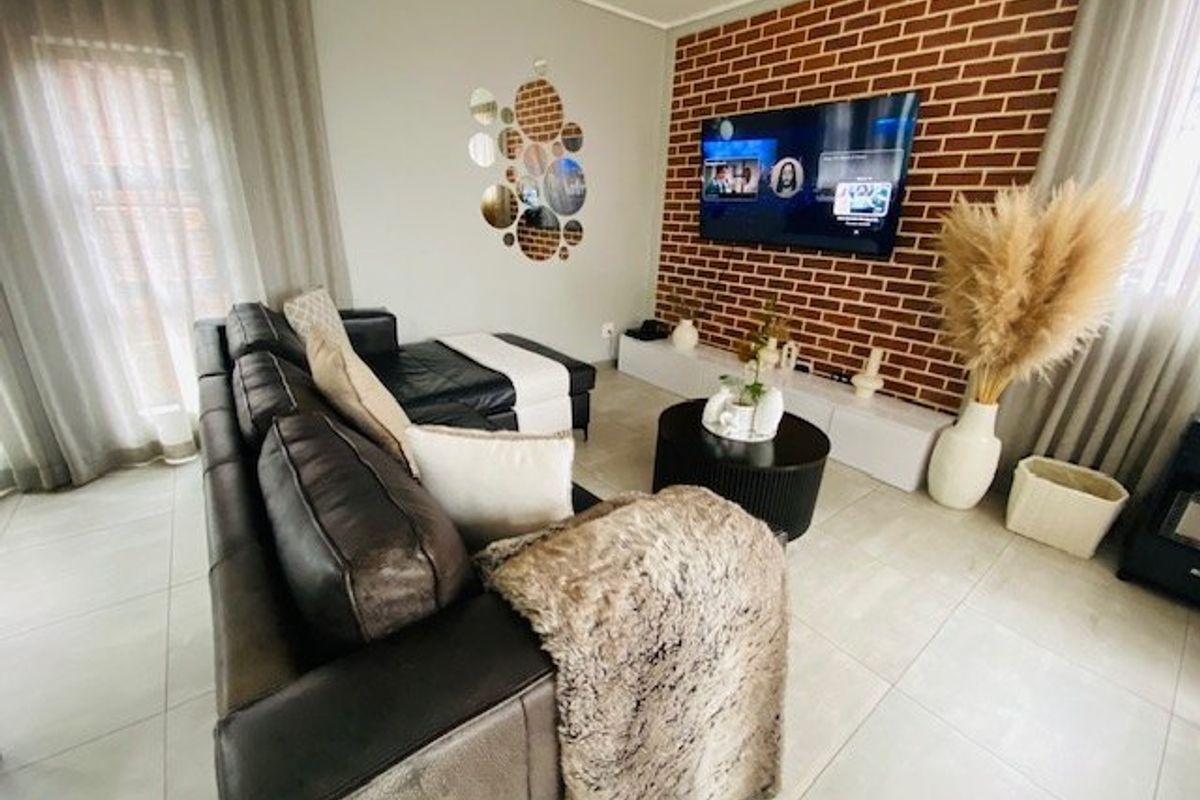 A well-maintained living room featuring a prominent exposed brick feature wall, neutral tiled flooring, and large windows with floor-to-ceiling curtains.