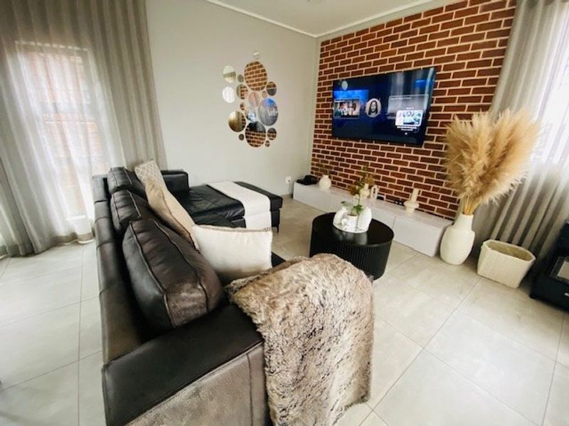 A well-maintained living room featuring a prominent exposed brick feature wall, neutral tiled flooring, and large windows with floor-to-ceiling curtains.