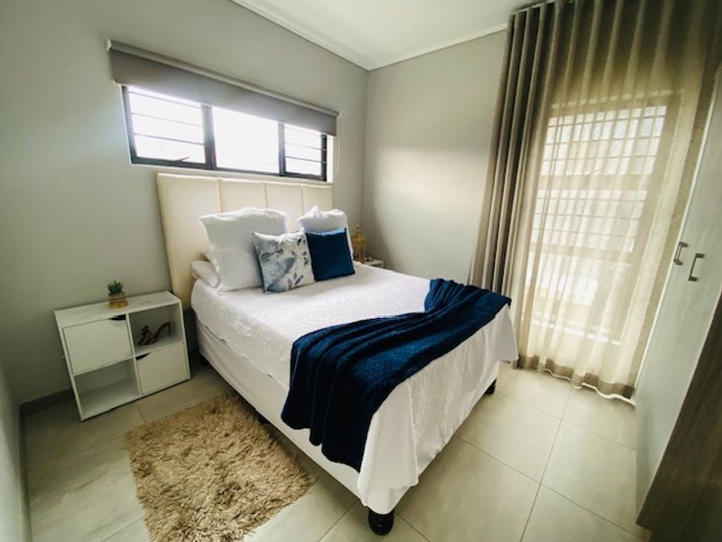 A well-maintained bedroom featuring neutral tiled flooring, a built-in wardrobe, and large windows with fitted blinds and curtains.