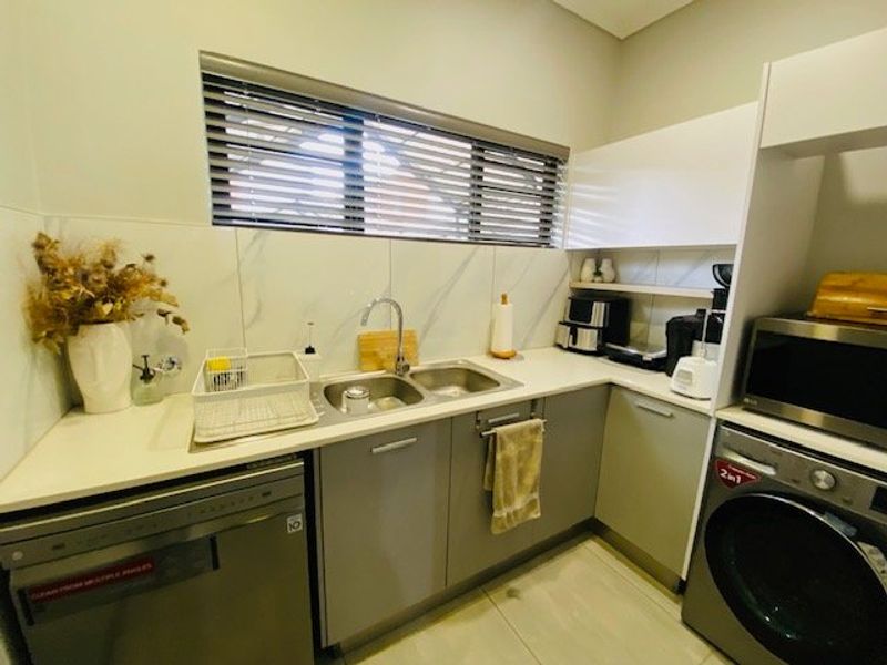 A functional scullery featuring modern grey cabinetry, stone countertops, a double basin sink, and tiled backsplash with integrated appliance space.