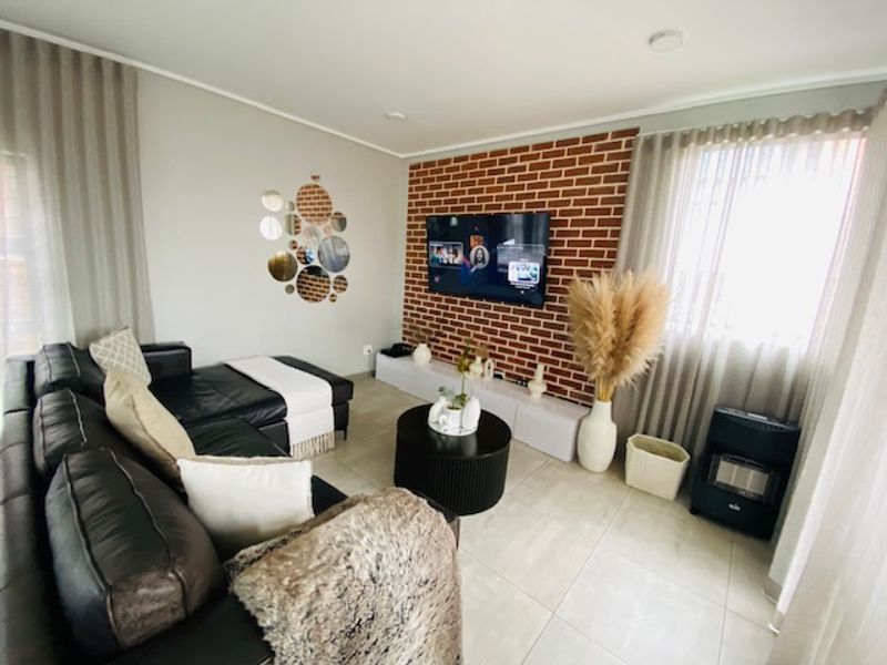 A well-maintained living room featuring a prominent exposed brick accent wall, neutral tiled flooring, and modern recessed lighting.