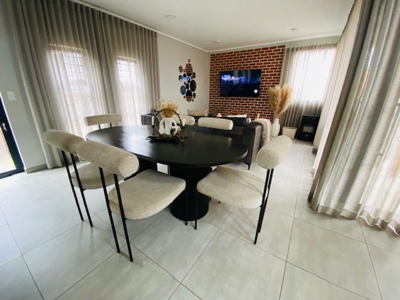 A bright and modern open-plan living and dining area featuring large windows, tiled flooring, and a striking exposed brick feature wall.