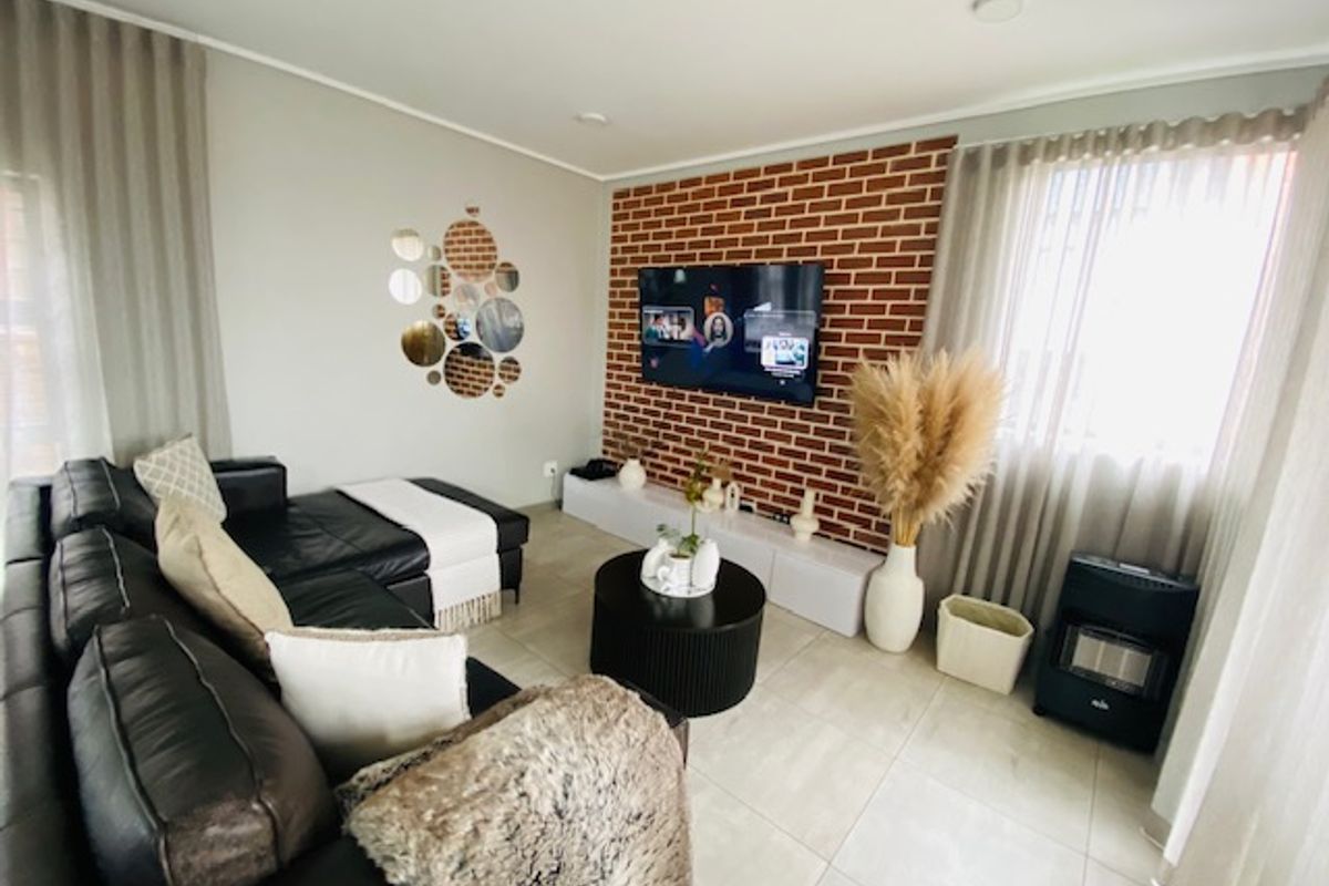 A well-maintained living room featuring a prominent exposed brick feature wall, neutral tiled flooring, and modern recessed lighting.