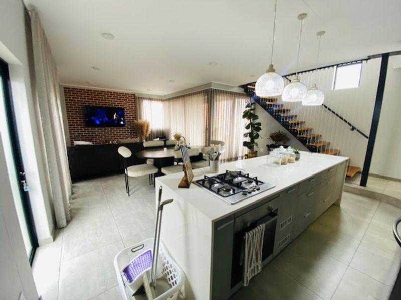 Modern open-plan living area featuring a central kitchen island with gas cooktop, exposed brick wall, and a contemporary staircase.