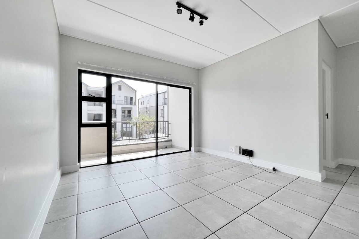 A bright, empty living room featuring neutral tiled flooring, modern track lighting, and a large sliding glass door leading to a balcony.