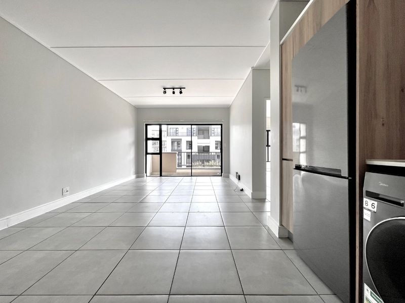 A bright, open-plan living and kitchen area featuring neutral tiled flooring, modern track lighting, and a sliding glass door leading to a balcony.