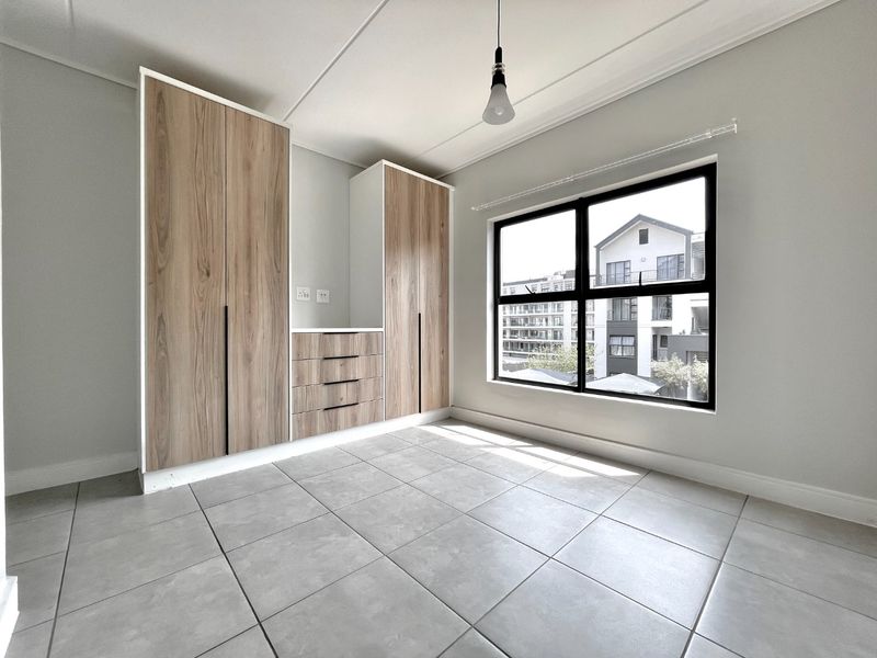 A bright, empty bedroom featuring modern wood-finish built-in cupboards, neutral tiled flooring, and large aluminium-framed windows providing natural light.