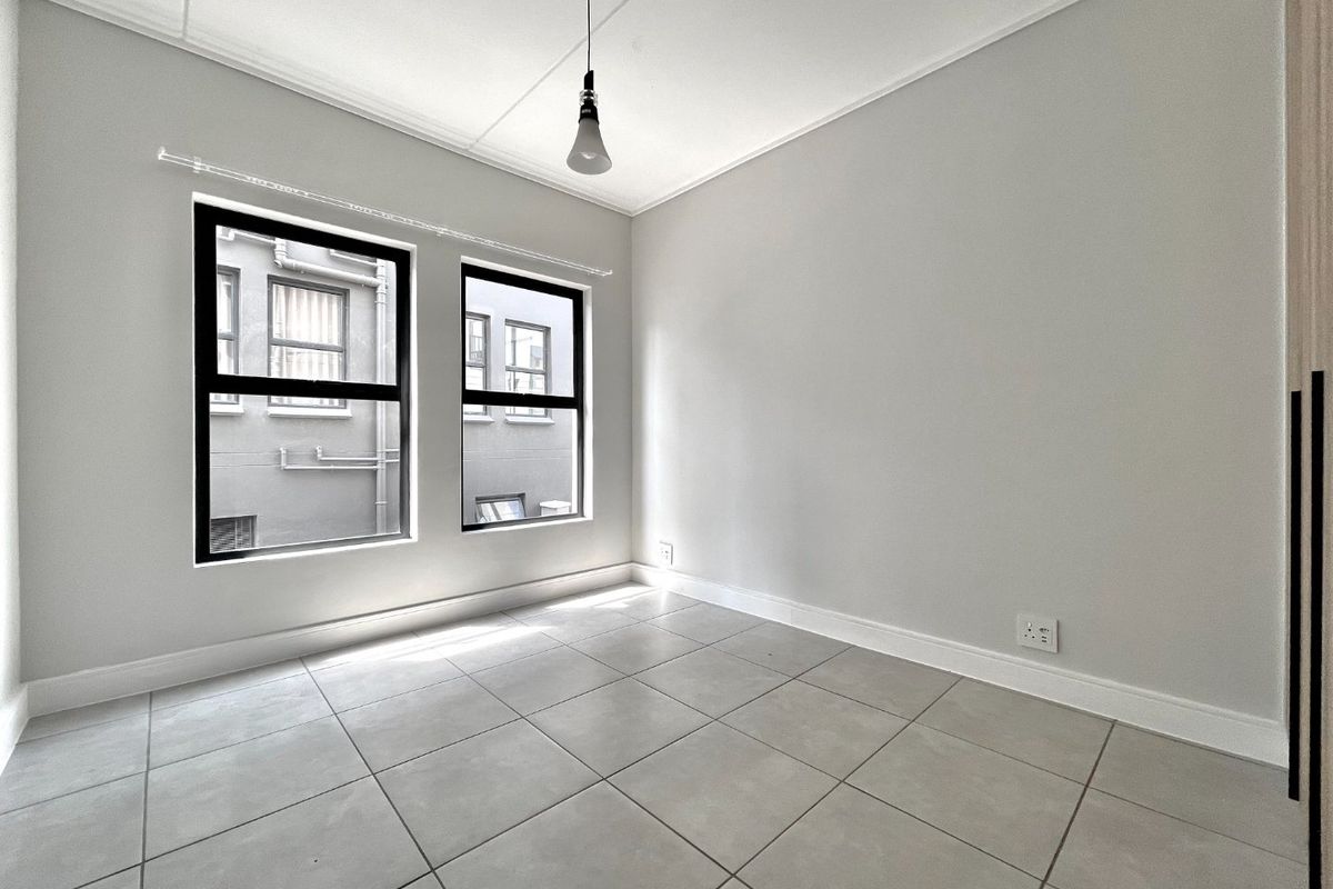 A bright, empty bedroom featuring neutral tiled flooring, modern black-framed windows, and a built-in wardrobe, offering a clean and versatile living space.