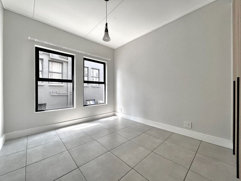 A bright, empty bedroom featuring neutral tiled flooring, modern black-framed windows, and a built-in wardrobe, offering a clean and versatile living space.