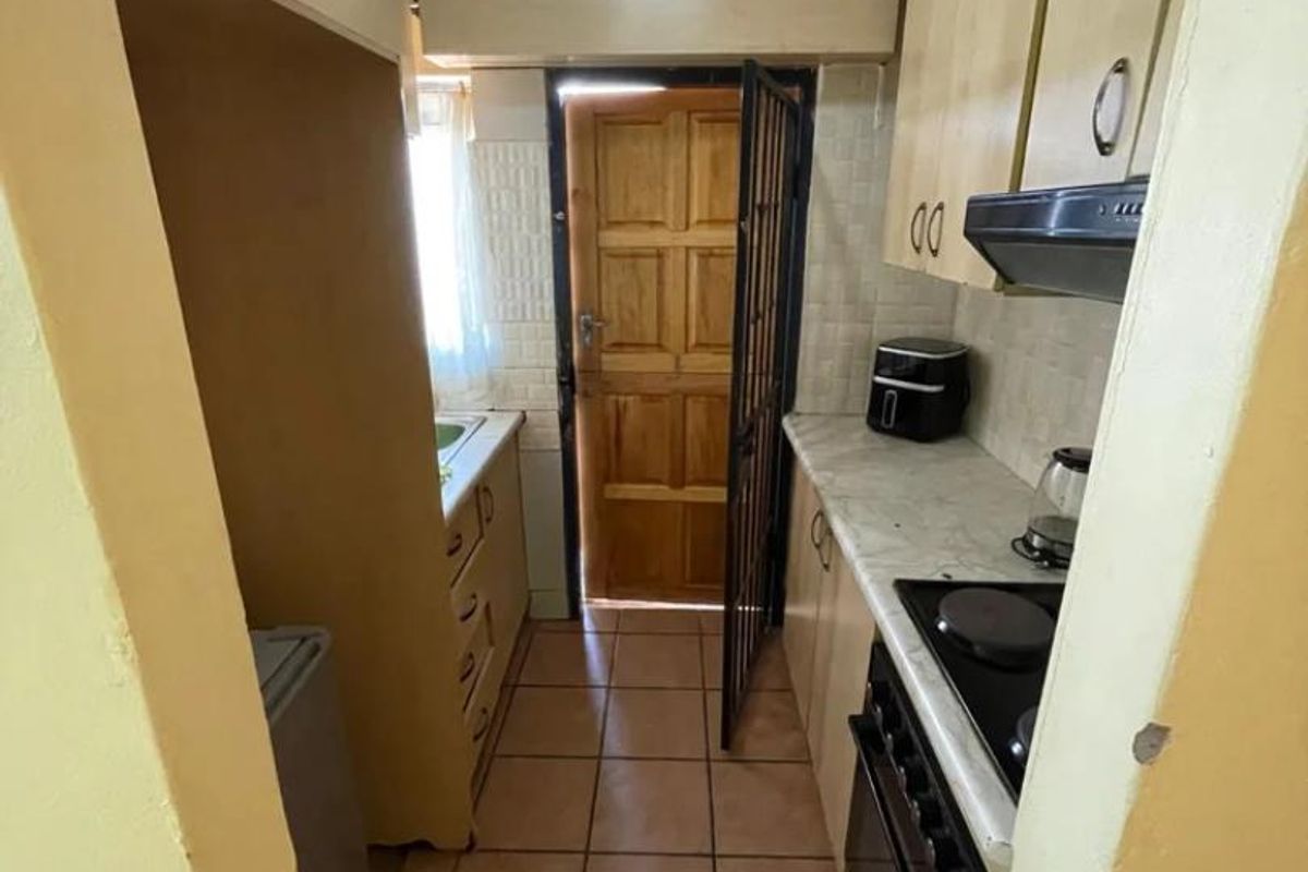 A compact galley-style kitchen featuring built-in cabinetry, electric stove with extractor fan, tiled flooring, and direct access to an exterior timber door.