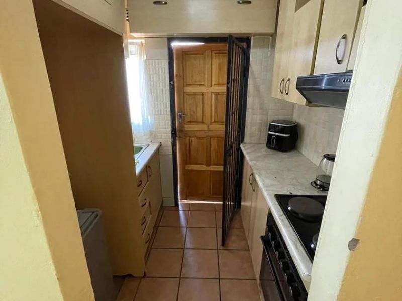 A compact galley-style kitchen featuring built-in cabinetry, electric stove with extractor fan, tiled flooring, and direct access to an exterior timber door.
