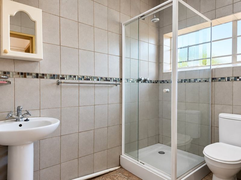 A functional bathroom featuring a pedestal basin, a glass-enclosed shower, and a toilet, finished with neutral wall tiles and a decorative mosaic border.
