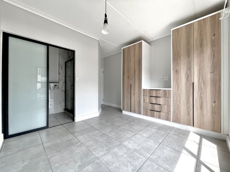 A bright, modern bedroom featuring neutral tiled flooring, ample built-in wooden cabinetry, and a sleek sliding glass door leading to an en-suite bathroom.