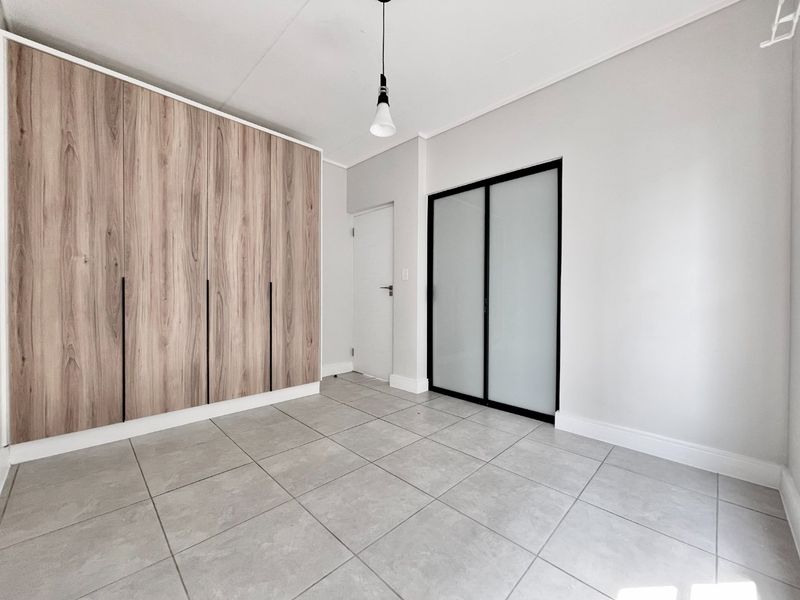 A bright, empty bedroom featuring modern wood-finish built-in cupboards, neutral tiled flooring, and a sleek frosted glass sliding door.