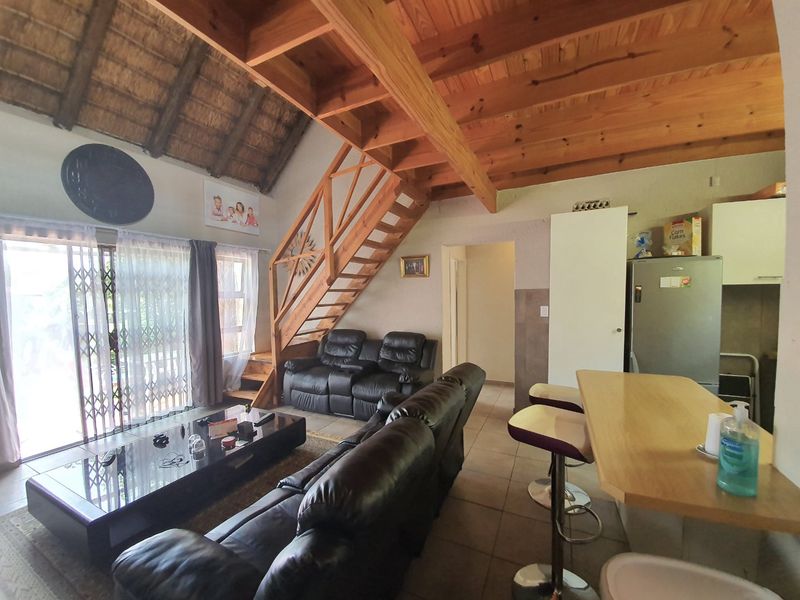 Open-plan living area featuring exposed timber rafters, a thatched ceiling, a wooden staircase, and a breakfast counter connecting to the kitchen space.
