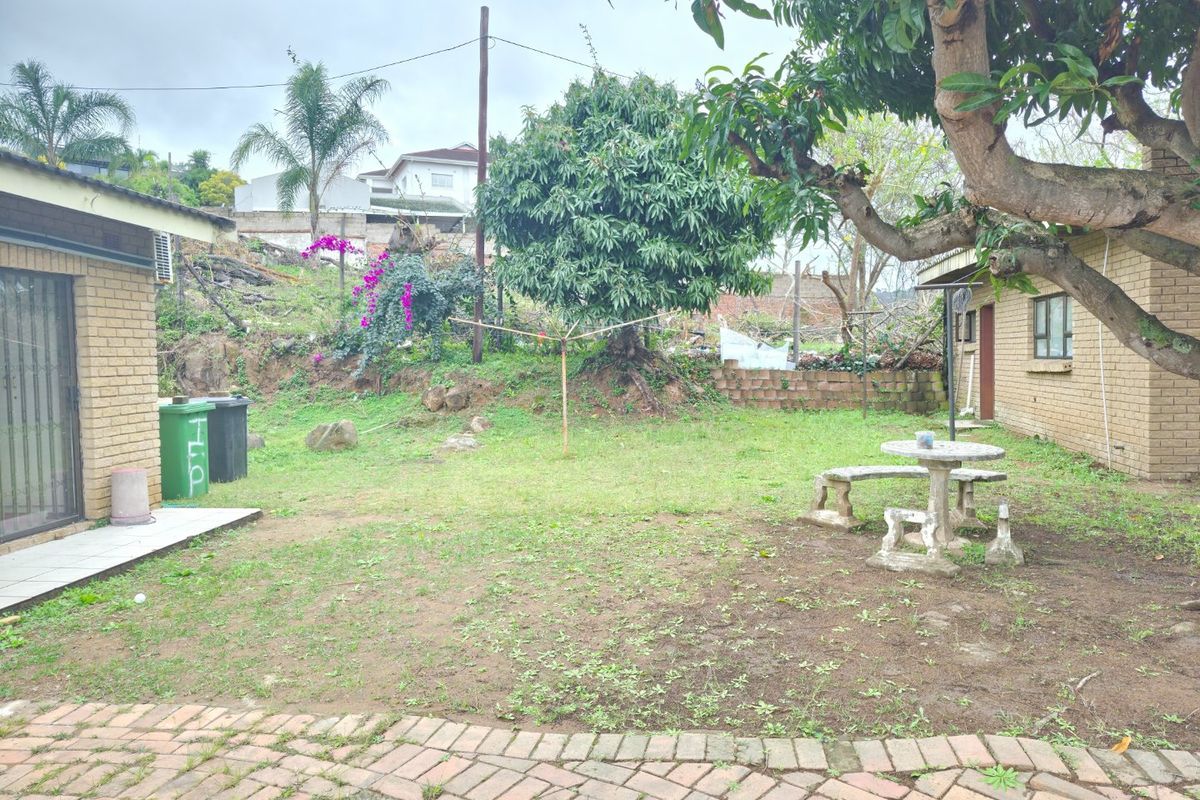 A rear garden area featuring a brick facade, paved patio space, and a sloped yard with established trees and retaining wall elements.