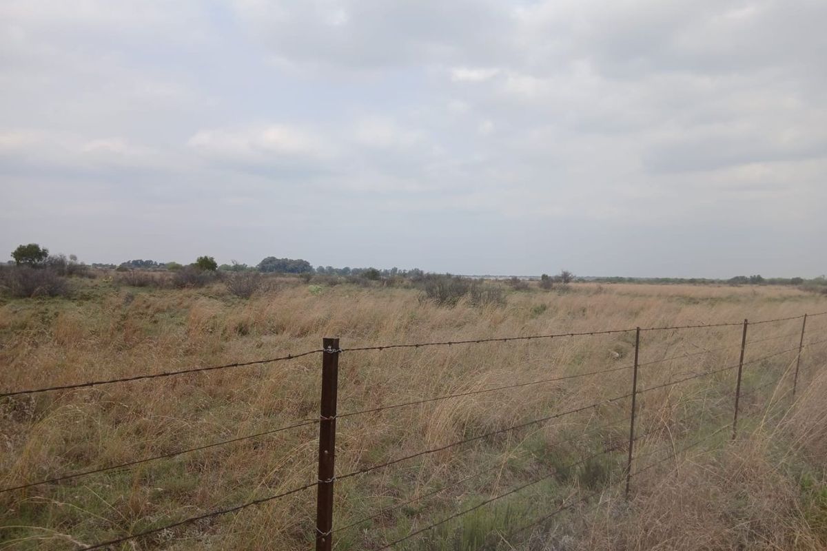 Expansive open farmland landscape featuring dry grass and a perimeter barbed wire fence under a cloudy sky.