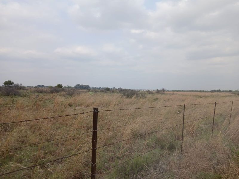 Expansive open farmland landscape featuring dry grass and a perimeter barbed wire fence under a cloudy sky.