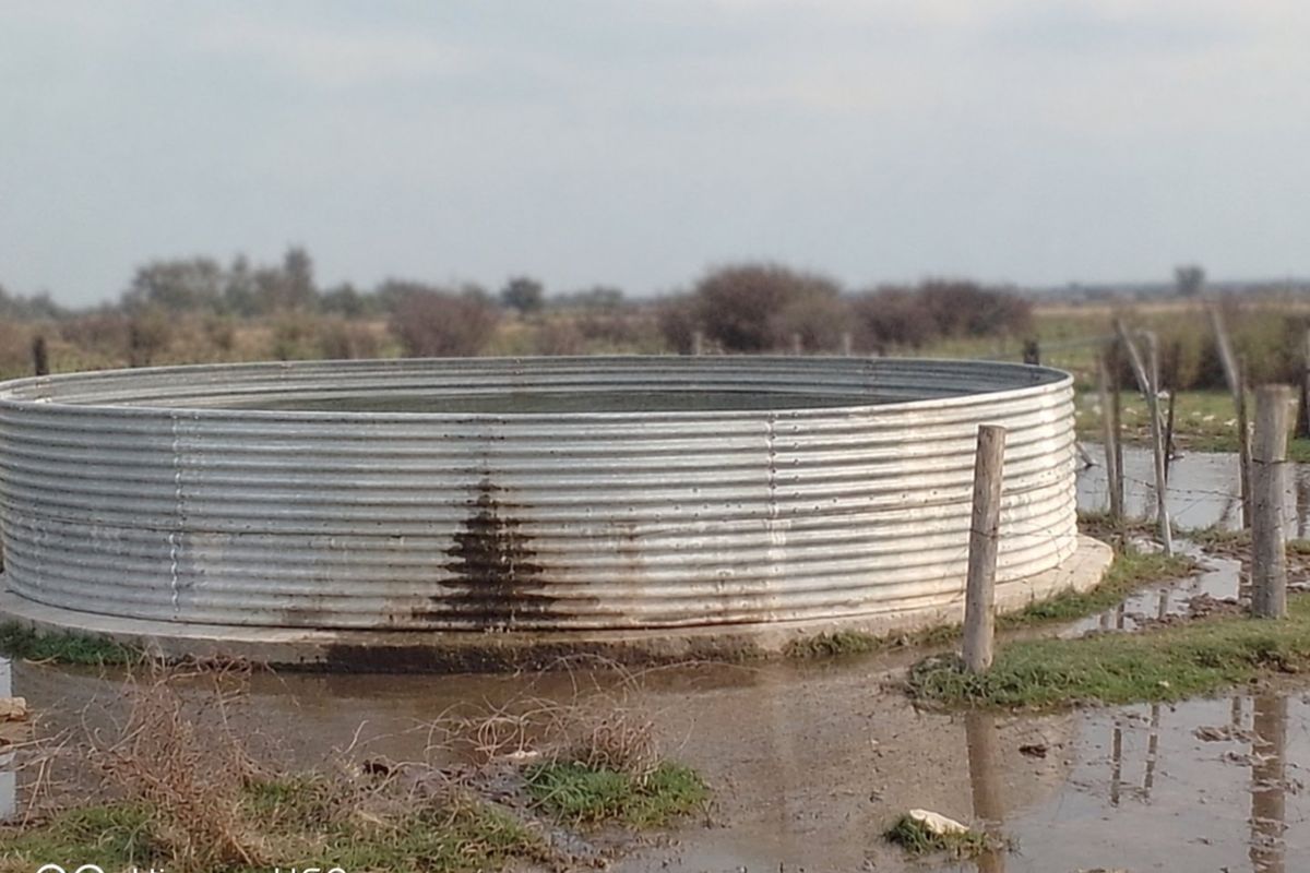 A corrugated metal water storage tank situated on a concrete base in a rural farm setting, showing signs of leakage and surrounding wet ground.