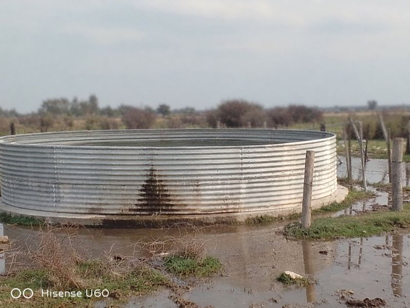 A corrugated metal water storage tank situated on a concrete base in a rural farm setting, showing signs of leakage and surrounding wet ground.