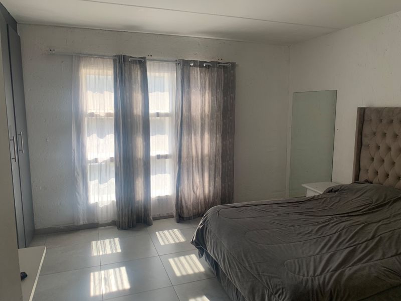 A bedroom featuring tiled flooring, a built-in wardrobe, and natural light through large windows. The room requires cosmetic updates to the wall finishes.