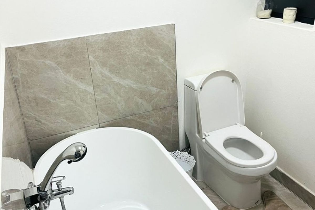 A clean, modern bathroom featuring a white freestanding bathtub with a handheld shower mixer, a toilet, and neutral-toned wall and floor tiling.