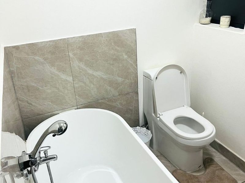 A clean, modern bathroom featuring a white freestanding bathtub with a handheld shower mixer, a toilet, and neutral-toned wall and floor tiling.