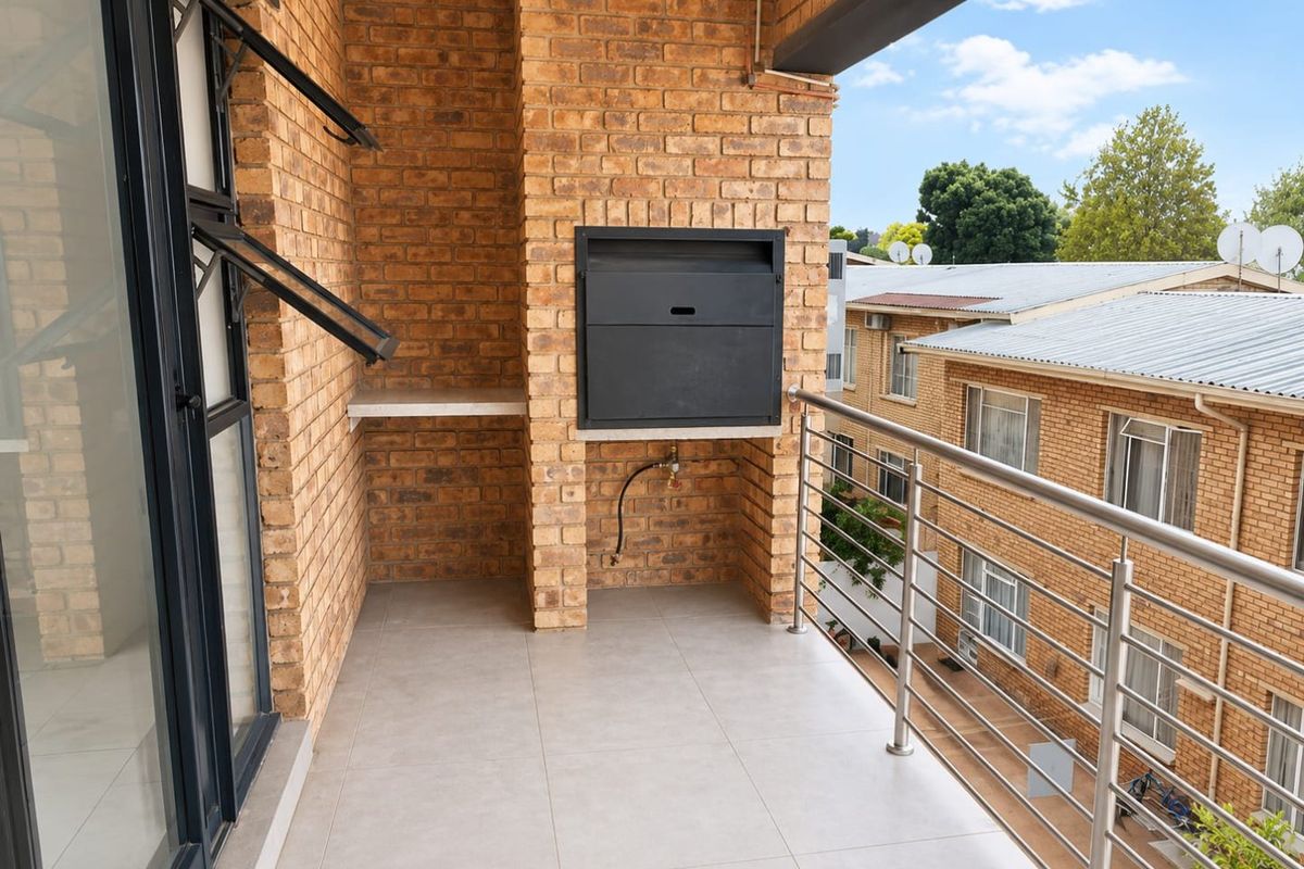 A clean, covered balcony featuring exposed brick walls, a built-in braai, and a modern stainless steel balustrade overlooking the complex.
