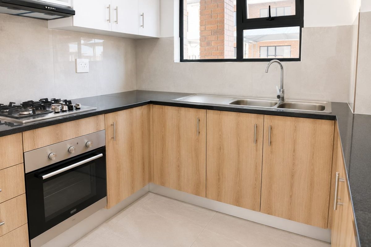Modern kitchen featuring light wood-finish cabinetry, granite countertops, stainless steel sink, gas hob, and built-in oven with a neutral tiled backsplash.