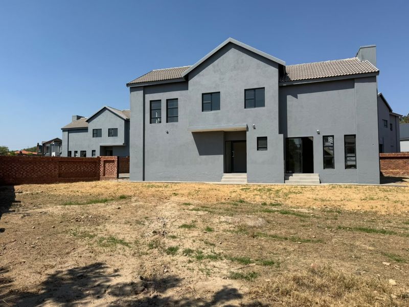 Rear view of a modern two-story grey house featuring a tiled roof, casement windows, and an expansive, undeveloped backyard with a brick boundary wall.