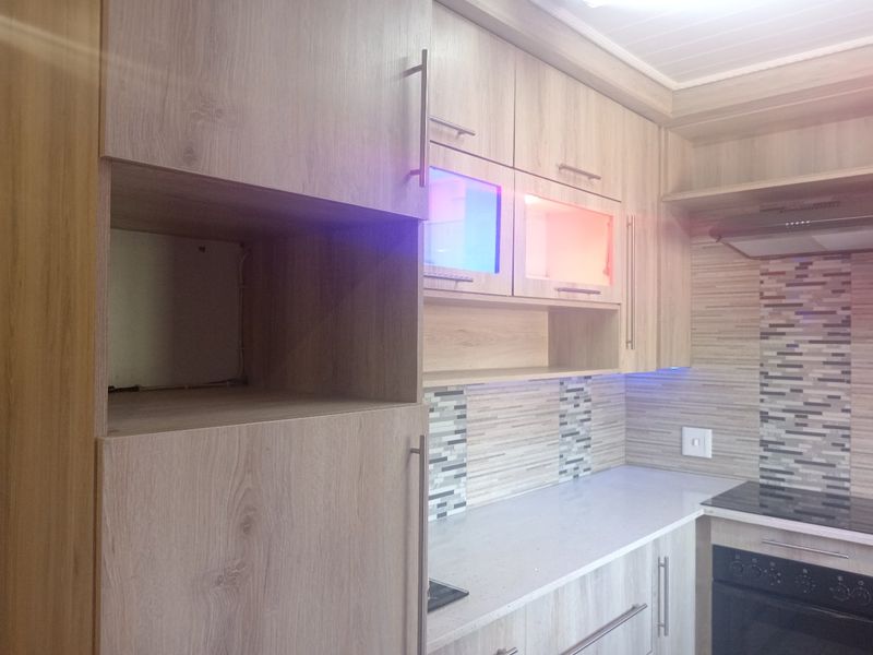 Modern kitchen featuring light wood-grain cabinetry, stone countertops, mosaic tile backsplash, and integrated appliances with ambient under-cabinet lighting.