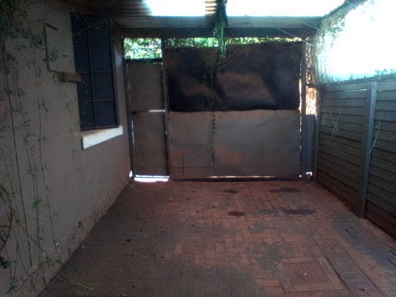 A covered outdoor garage or utility area featuring brick paving, timber fencing, and a metal gate, currently showing signs of wear and maintenance needs.