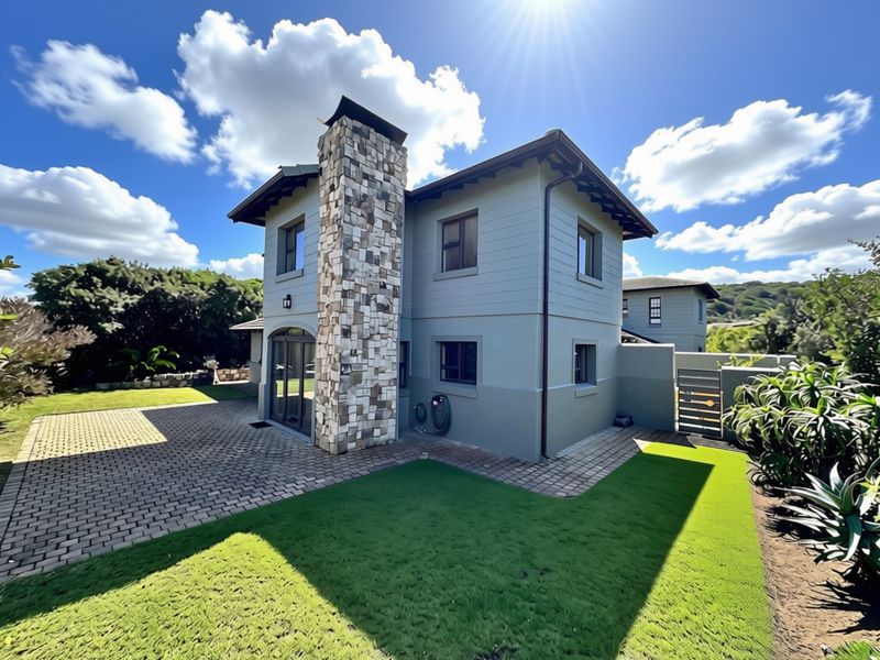 Rear view of a modern two-story apartment featuring a stone chimney, paved patio area, and well-maintained artificial lawn.